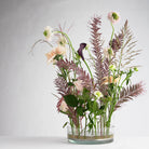 A glass vase, enhanced with a flower frog ring, holds a bouquet of beautiful flowers and leaves, featuring delicate pink blooms, a dark purple lily, and feathery pink foliage, all arranged against a plain white background.