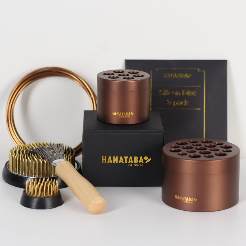 A set of Hanataba Original flower arrangement tools, featuring two muddy brown floral holders, a larger kenzan (70mm), a smaller kenzan (34mm) and a kenzan rake brush with a wooden handle. The items are arranged on a black box with golden Hanataba logo lettering.