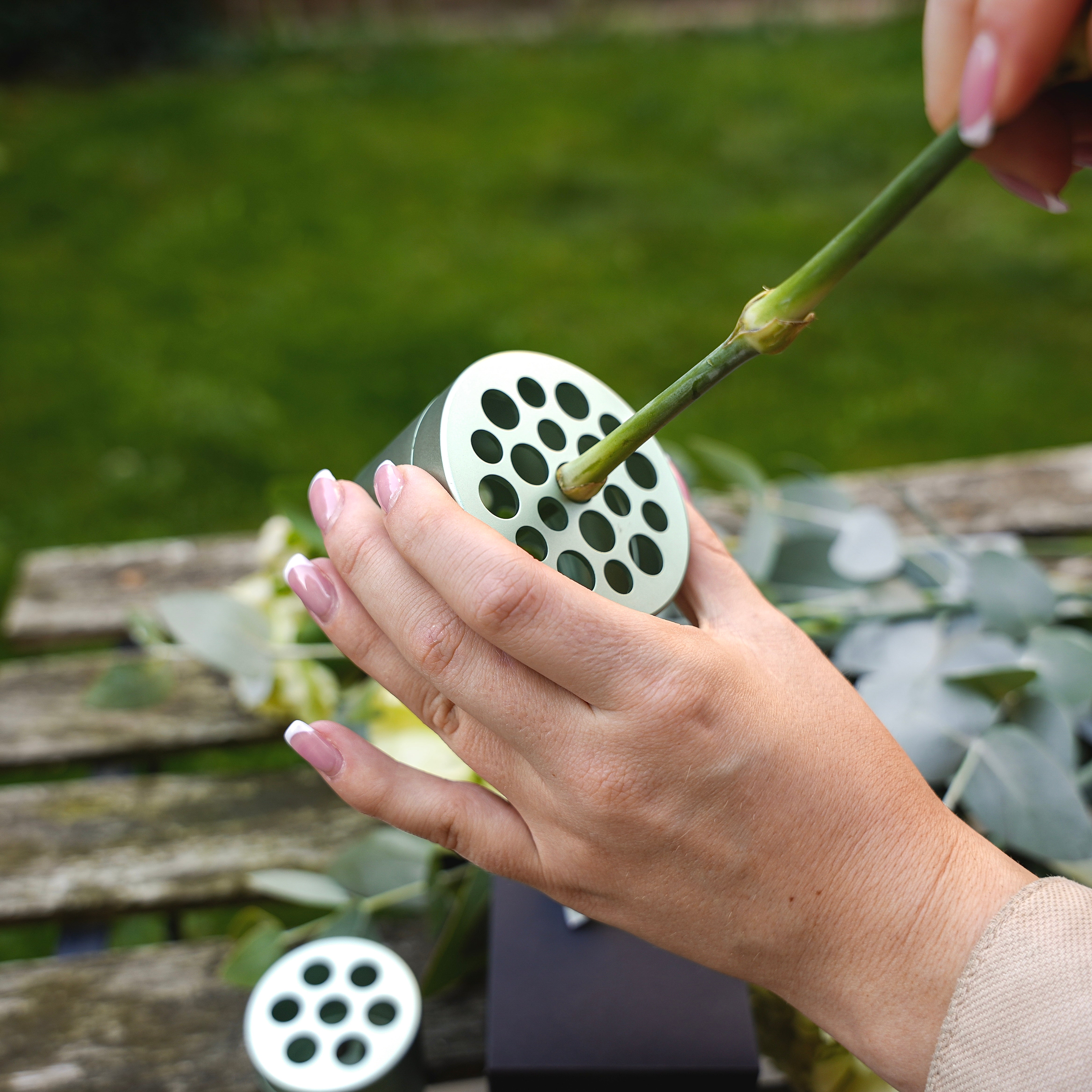 A person with pink nail polish arranging eucalyptus and flowers by inserting a stem into a Hanataba Matcha Green bouquet twister tool over a wooden table in the garden.