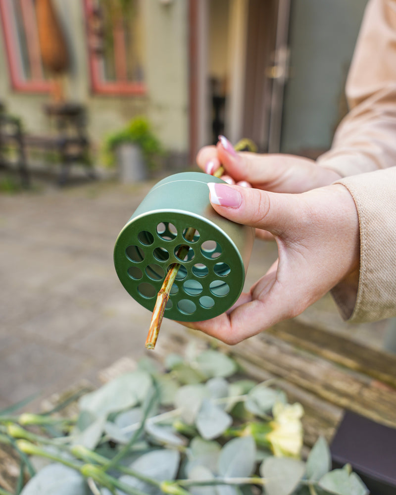 Osoba trzymająca żabę kwiatową Hanataba Ivy Green z łodygą kwiatu włożoną przez jeden z otworów. Liście eukaliptusa i inna zieleń są rozrzucone na powierzchni poniżej. Tło przedstawia plener.