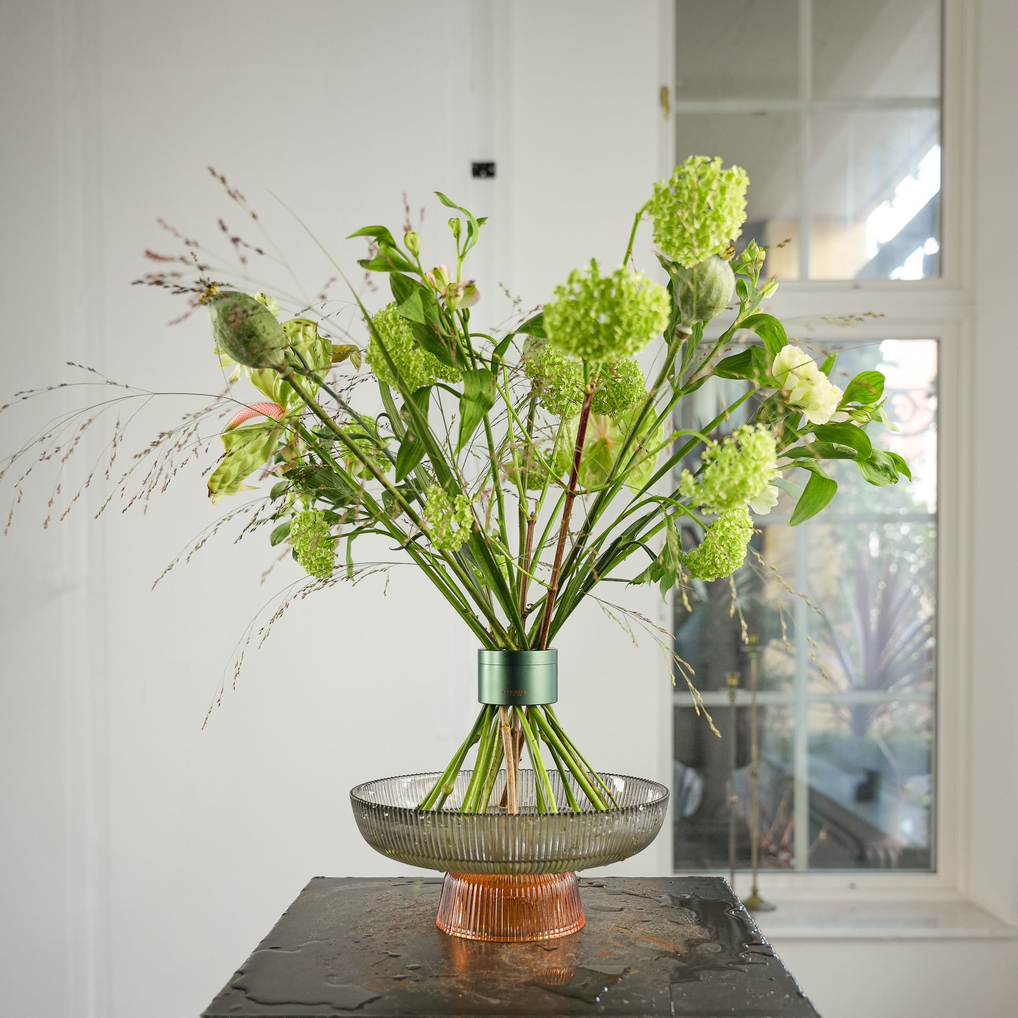 Stunning bouquet of grass and viburnum flowers arranged in a Hanataba Ivy Green tool, displayed in a glass tray with water on a black table by a window in a bright white room.