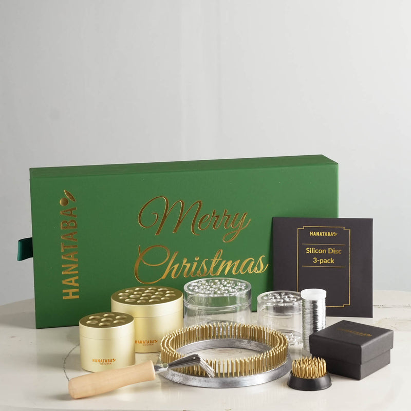 A Christmas gift set in a green box with "Merry Christmas" in gold, featuring flower arranging essentials such as a 120mm kenzan ring, a 34mm flower frog and four Hanataba flower twisters. The products are displayed elegantly on a light surface.