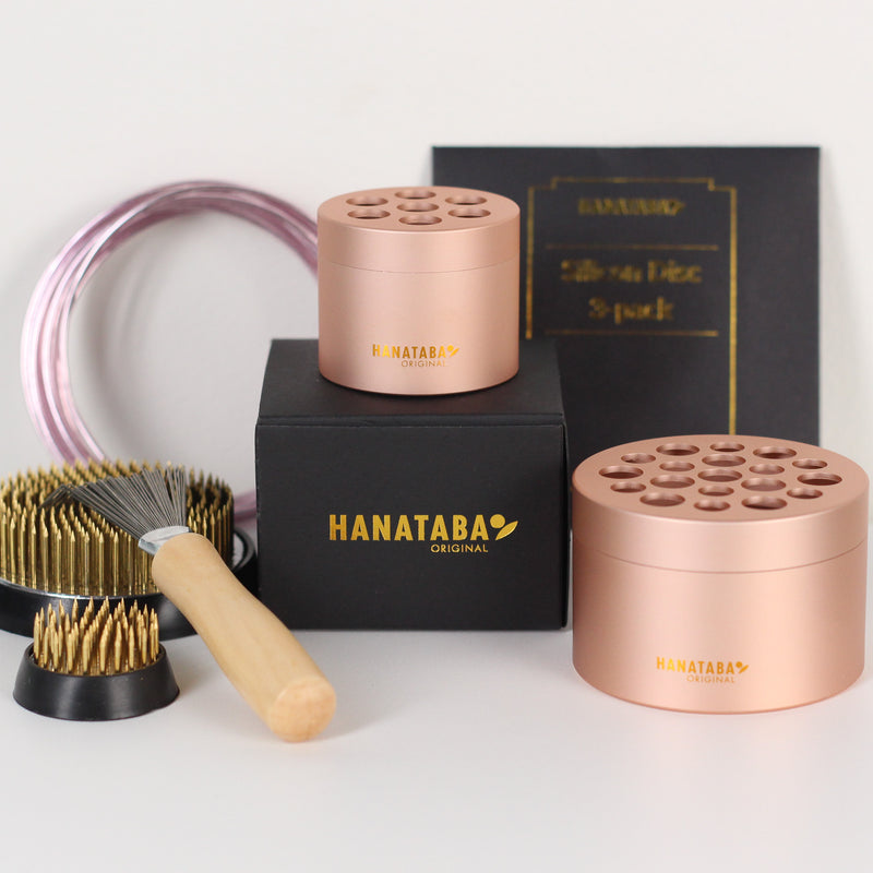 Zestaw oryginalnych narzędzi do układania kwiatów Hanataba, zawierający dwa stojaki na kwiaty Hanataba Pink Champagne, większy kenzan (70 mm), mniejszy kenzan (34 mm) i szczotkę do grabienia kenzan z drewnianą rączką. Przedmioty są umieszczone w czarnym pudełku ze złotym logo Hanataba.