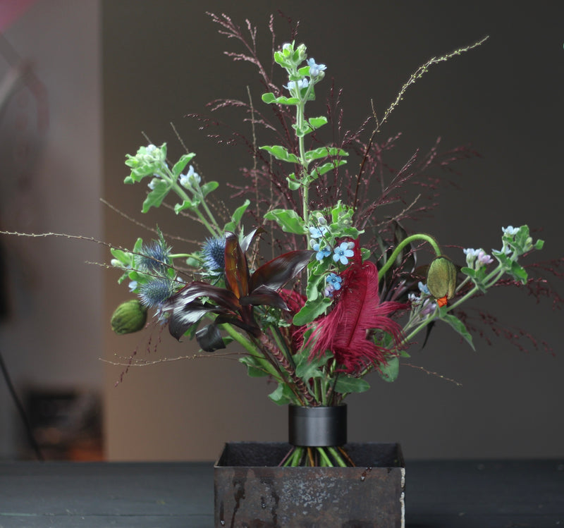 Aranżacja Ikebana zawierająca różnorodne kwiaty i liście zabezpieczone uchwytem Hanataba z czarnymi spiralnymi łodygami.