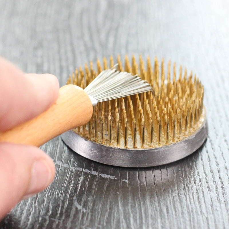 Close-up of a hand using a Kenzan cleaning brush with a wooden handle to maintain the golden needles of a flower frog, ensuring precision and care in ikebana arrangements.