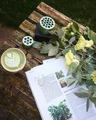 Eucalyptus and a cup of matcha tea next to the Hanataba bouquet twister tool on a wooden table in a garden setting.