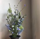 An eclectic mix of blue and white wildflowers with delicate greenery, displayed in a dark bowl on a kenzan ring, against a neutral background.